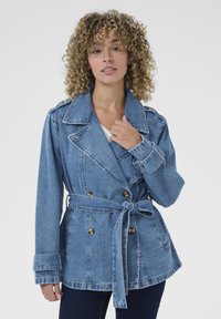 Ženska s kodrastimi lasmi, oblečena v modro denim jakno z beltom in temne hlače, drži ovratnik jakne na enobarvni beli podlagi.