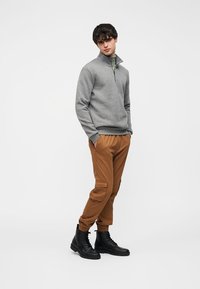 Sweat-shirt gris à col zippé, pantalon cargo marron, et bottes à lacets noires. Tissu lisse, coupe décontractée, poches visibles sur le pantalon, et poignets côtelés.