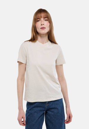 Jeune femme aux cheveux roux raides portant un t-shirt beige uni à manches courtes et un jean bleu, debout devant un fond blanc.