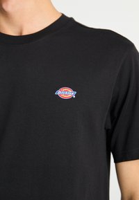 Black cotton t-shirt with short sleeves and a crew neck; features a small, colorful "Dickies" logo on the upper left chest.