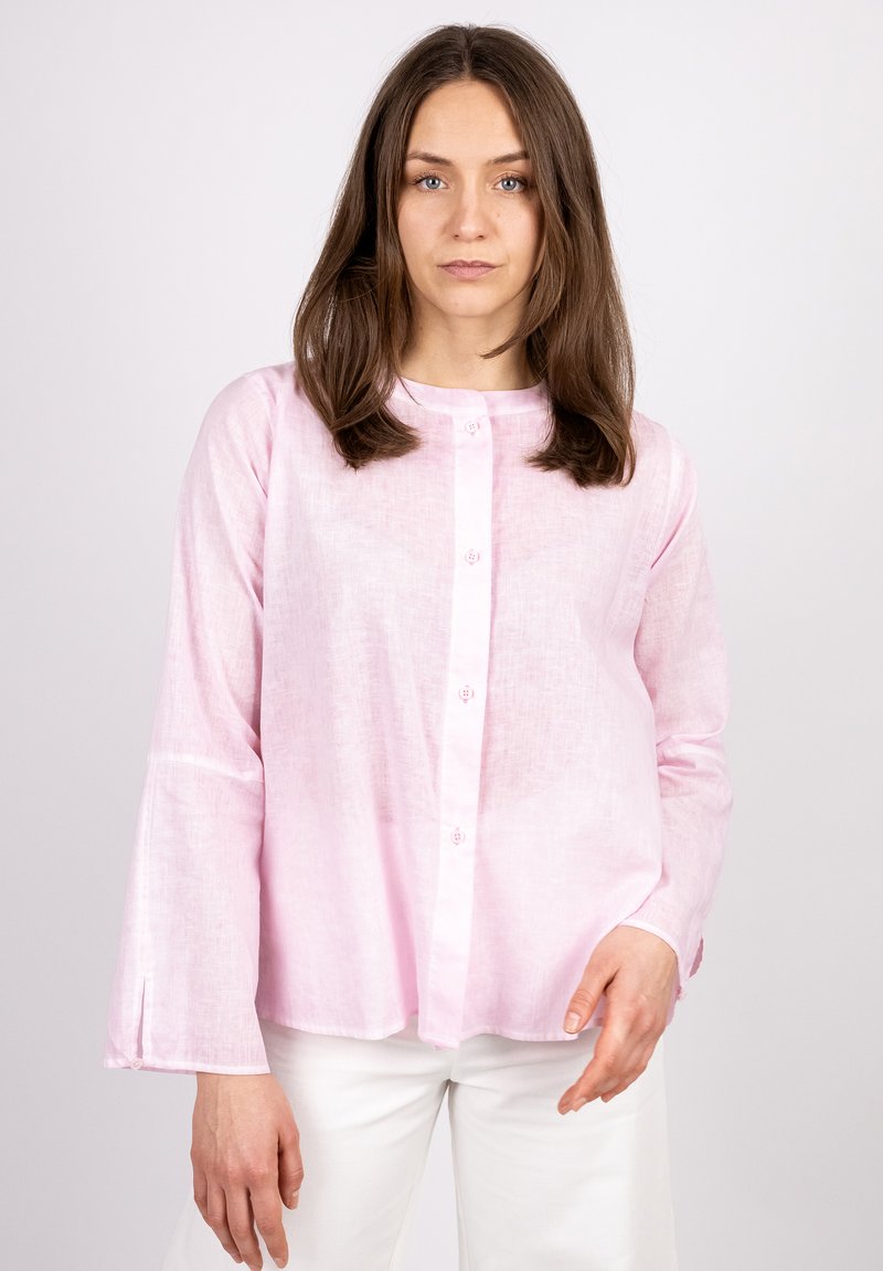 Helle rosa Bluse mit langen Ärmeln, Rundhalsausschnitt, Knöpfen an der Brust und seitlichen Schlitzen. Hergestellt aus einem strukturierten, leichten Stoff.