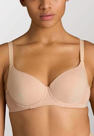 Donna che indossa un reggiseno beige con ferretto e spalline regolabili su uno sfondo chiaro uniforme.