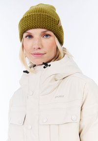 Vrouw met blond haar, gekleed in een olijfgroene gebreide muts en een beige winterjas met capuchon en knoopzakken aan de borst.