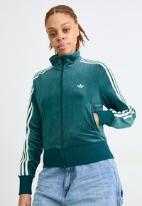 Pessoa jovem com cabelo trançado veste uma jaqueta zip-up Adidas teal com listras brancas e jeans azul claro, em pé contra um fundo branco.