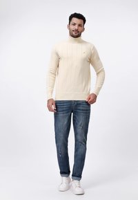 Pull à col roulé côtelé crème associé à un jean bleu déchiré et des baskets blanches, avec un logo minimaliste sur la poitrine.