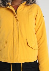 Personne portant une veste à capuche jaune avec bordure en fourrure, fermeture éclair à l'avant, poches à rabat, et pantalon noir visible de la taille vers le bas.