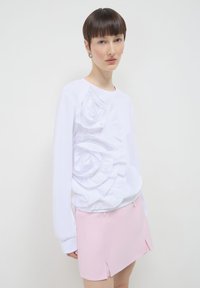 Felpa bianca a maniche lunghe con rose in tessuto tridimensionale sul lato sinistro, abbinata a una mini gonna rosa chiaro con fessure laterali.