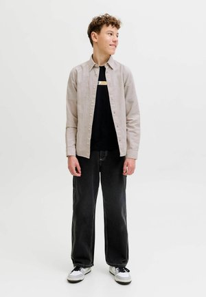 Mladý človek s kučeravými vlasmi, oblečený v béžovej košeli na gombíky cez čierne tričko s potlačou, čiernych širokých džínsach a bielych teniskách, pozerajúci sa doprava.