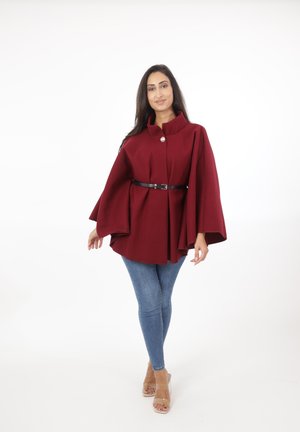 Burgundyröd cape med hög krage, med knappstängning fram och ett svart bälte i midjan, som bärs över ljusblå jeans.