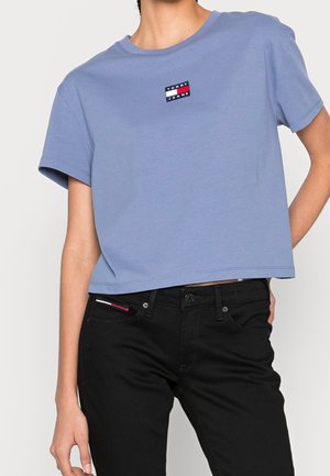 Persoon draagt een kort lichtblauw Tommy Jeans t-shirt en zwarte jeans, waarbij een ingestoken kledingstuk de rode, witte en blauwe bies van het merk toont.