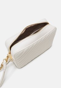 Bílá kabelka z umělé kůže s prošívaným vzorem, zlatými kovovými doplňky a zapínáním na zip. Vnitřek je vyložený vzorovanou látkou. Součástí je nastavitelný popruh.