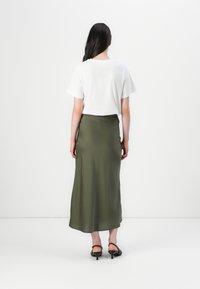 En person med långt hår står vänd bort, klädd i en vit t-shirt och en lång olivgrön kjol, med en minimalistisk bakgrund.