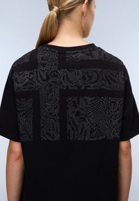 Černé bavlněné tričko s texturovaným grafickým designem ve stříbrných tečkách na zadní straně, volného střihu a krátkými rukávy.