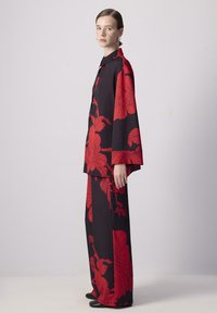 Schwarzes Outfit mit großem rotem Blumenmuster, bestehend aus einer knopfbaren Bluse mit weiten Ärmeln und passenden, locker sitzenden Hosen. Glatte Stoffstruktur.