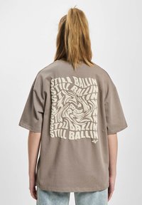 Gray oversized t-shirt with short sleeves, featuring a swirling white “STILL BALLIN” text design on the back. Soft fabric texture.