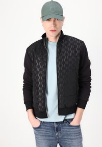 Μαύρο μπόμμερ μπουφάν με patterned μπροστινό μέρος, μαύρους μανίκους και φερμουάρ. Ανοιχτό μπλε t-shirt από κάτω. Γκρι καπέλο με λογότυπο. Τζιν παντελόνι.