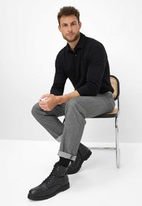 Zwart langemouwen polo, grijze textuurbroek en zwarte veterschoenen. Model zit op een lichte stoel met gevlochten zitting. Minimale achtergrond.