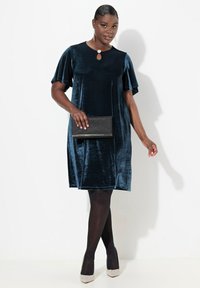 Laivansinis samettimekko, jossa on lyhyet leijonahihnat ja avonainen kaula-aukko. Yhdistetty mustaan clutch-laukkuun ja kimaltaviin korkokenkiin.