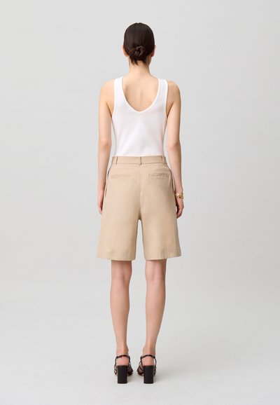 Mujer de espaldas, vestida con una camiseta blanca sin mangas, pantalones cortos beige hasta la rodilla, sandalias negras de tacón y una pulsera dorada.