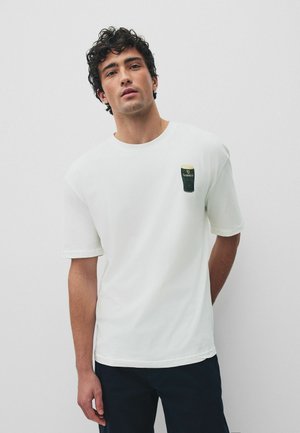 Vyras su garbanotais plaukais, dėvintis baltą marškinėlį su mažo Guinness alaus stiklo grafika kairėje krūtinės pusėje, stovintis priešais paprastą foną.