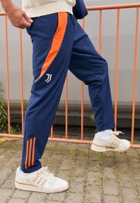 Pantaloni da jogging blu navy con strisce arancioni ai lati, dotati di un logo bianco e tre strisce arancioni alla caviglia. Indossati con sneakers beige.
