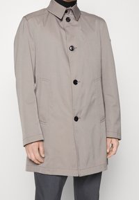 Cappotto impermeabile beige con colletto, chiusura frontale con bottoni, due tasche laterali e maniche lunghe. Tessuto liscio con accenti neri a contrasto.