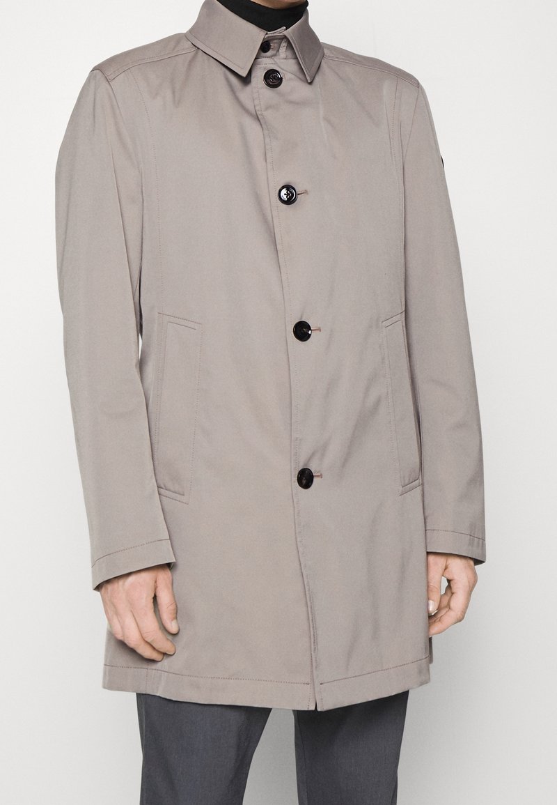 Cappotto impermeabile beige con colletto, chiusura frontale con bottoni, due tasche laterali e maniche lunghe. Tessuto liscio con accenti neri a contrasto.