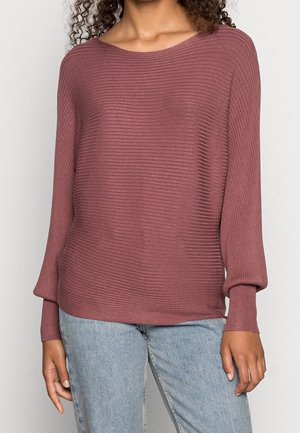 Kobieta w długim rękawie, prążkowanym, wrzosowym swetrze z mankietami oraz jasnoniebieskich dżinsach, stojąca na jednolitym tle.