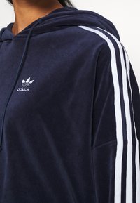 Mörkblå hoodie i mjukt tyg, med en framficka, huva med dragsko och vita ränder längs ärmarna samt en broderad logotyp.