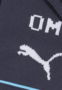 Tume sinine kootud kangas, millel on valge "OM" tekst ja valge Puma logo, kerge sinise horisontaalse aktsentjoonega.