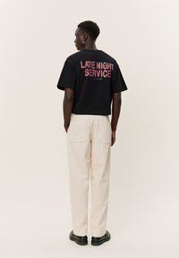 Oseba v črni majici z napisom »LATE NIGHT SERVICE« na hrbtu, kremnih hlačah in črnih čevljih, stoji pred enobarvnim ozadjem.