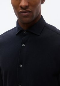 Granatowa koszulka polo z fakturowanej tkaniny, z ostrym kołnierzykiem i dwoma srebrnymi guzikami. Zbliżenie na górną część koszulki.