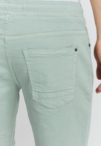 Pantalonii verde deschis au un material texturat, buzunare posterioare cu clapete înclinate și detalii contrastante din accesorii negre.