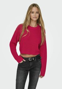 Maglione corto rosa a maglia con texture a coste e maniche lunghe, abbinato a jeans neri a vita alta e una cintura nera.