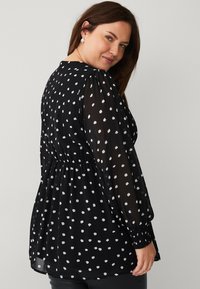 Zwarte blouse met witte stippen, doorschijnende stof, ronde onderkant, lange pofmouwen en elastische manchetten. Geverzameld bij de taille.