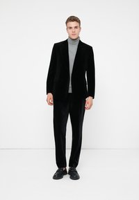 Blazer di velluto nero sopra un dolcevita grigio, abbinato a pantaloni di raso neri e scarpe slip-on nere. Texture liscia e vestibilità su misura.
