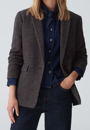 Vrouw draagt een grijze blazer met textuur over een donkerblauw hemd met knopen en een strikje bij de hals, gecombineerd met een donkere spijkerbroek, met één hand in de zak.
