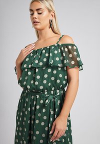 Vihreä pilkullinen off-the-shoulder-mekko, jossa on röyhelösateet, kireä vyötärö ja yhteensopiva vyö. Teksturoitu kangas, jossa on pehmeä ja virtaava muotoilu.