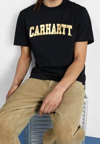 Černé bavlněné tričko s velkým, výrazným zlatým nápisem "CARHARTT" přes hruď. V kombinaci s béžovými kordovými kalhotami.