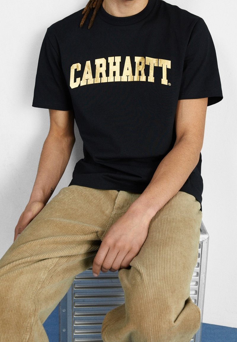 Černé bavlněné tričko s velkým, výrazným zlatým nápisem "CARHARTT" přes hruď. V kombinaci s béžovými kordovými kalhotami.