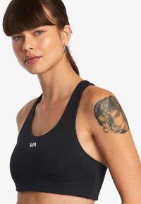 Sujetador deportivo negro hecho de tela elástica; cuenta con un escote en forma de scoop y tirantes gruesos, con un pequeño logo blanco en la parte delantera.