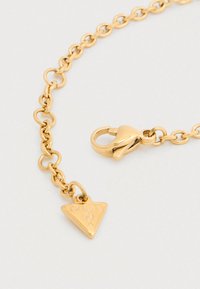 Βραχιόλι αλυσίδας σε χρυσή απόχρωση με οβάλ κρίκους και τριγωνικό μενταγιόν χαραγμένο με τη λέξη "GUESS", κλειστό με κούμπωμα.