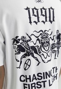 Hvid t-shirt med sort grafisk design, der inkluderer elementer som figurer, en maske og teksten "1990 CHASING THE FIRST LIGHT". Blødt bomulds materiale.