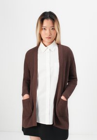 Young woman with straight blonde hair wears a white buttoned shirt, brown cardigan with pockets, and black skirt, facing the camera.