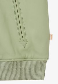 Hellgrüner Lederjackenärmel mit diagonaler, gesteppten Taschenverzierung, geripptem Strickbündchen und kleinem beigem Stoffmarkenetikett an der Seite.