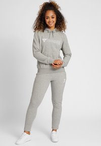 Ljusgrå hoodie och matchande joggers tillverkade av mjukt material, med en vit logotyp på bröstet och låret, samt ribbade ärmslut och midja.