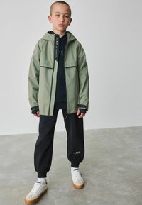 Lichtgroene regenjas met capuchon, twee voorzakken, zwarte accenten, gelaagd over een zwarte hoodie, gecombineerd met zwarte trainingsbroek en sneakers.
