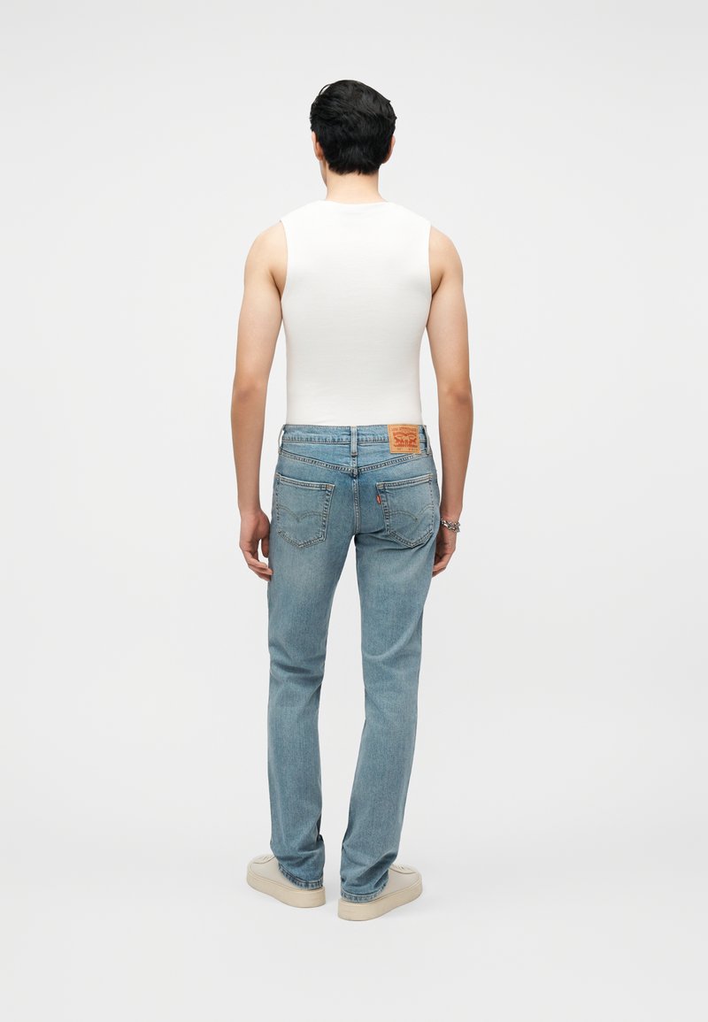 Mann trägt weißes ärmelloses Hemd, hellblaue Jeans mit Lederpatch, beige Turnschuhe, steht mit dem Rücken zur Kamera vor einfarbigem Hintergrund.