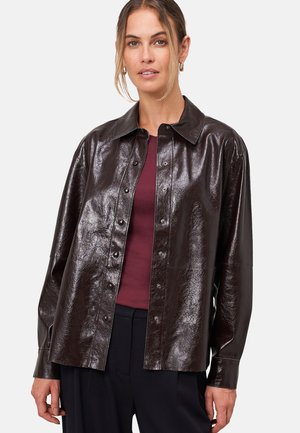 Une femme portant une chemise en cuir verni marron foncé à boutons sur un haut bordeaux et un pantalon noir, avec des boucles d'oreilles en or et les cheveux attachés lâchement.