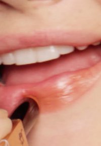 Primo piano di labbra con una finitura lucida, che mostrano una tonalità di rosa caldo. Un applicatore marrone viene applicato sul labbro inferiore.